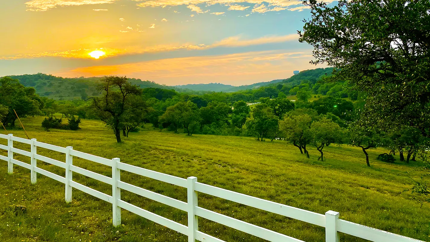 A white wooden fence running along a lush green pasture with rolling hills and scattered trees under a glowing sunset.