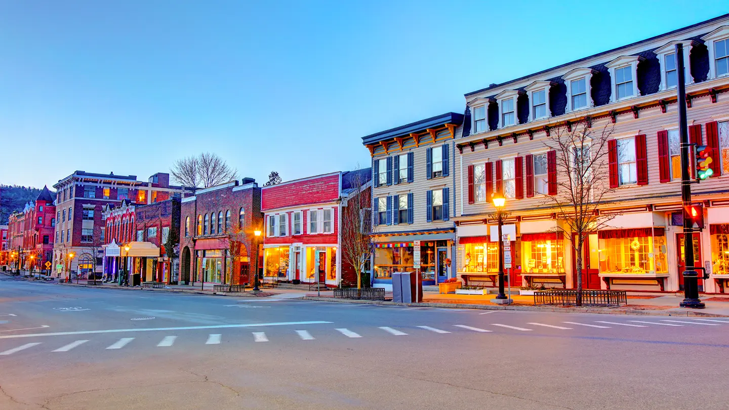 A quiet small-town main street at twilight, lined with colorful historic multi-story buildings and warm glowing storefront windows.