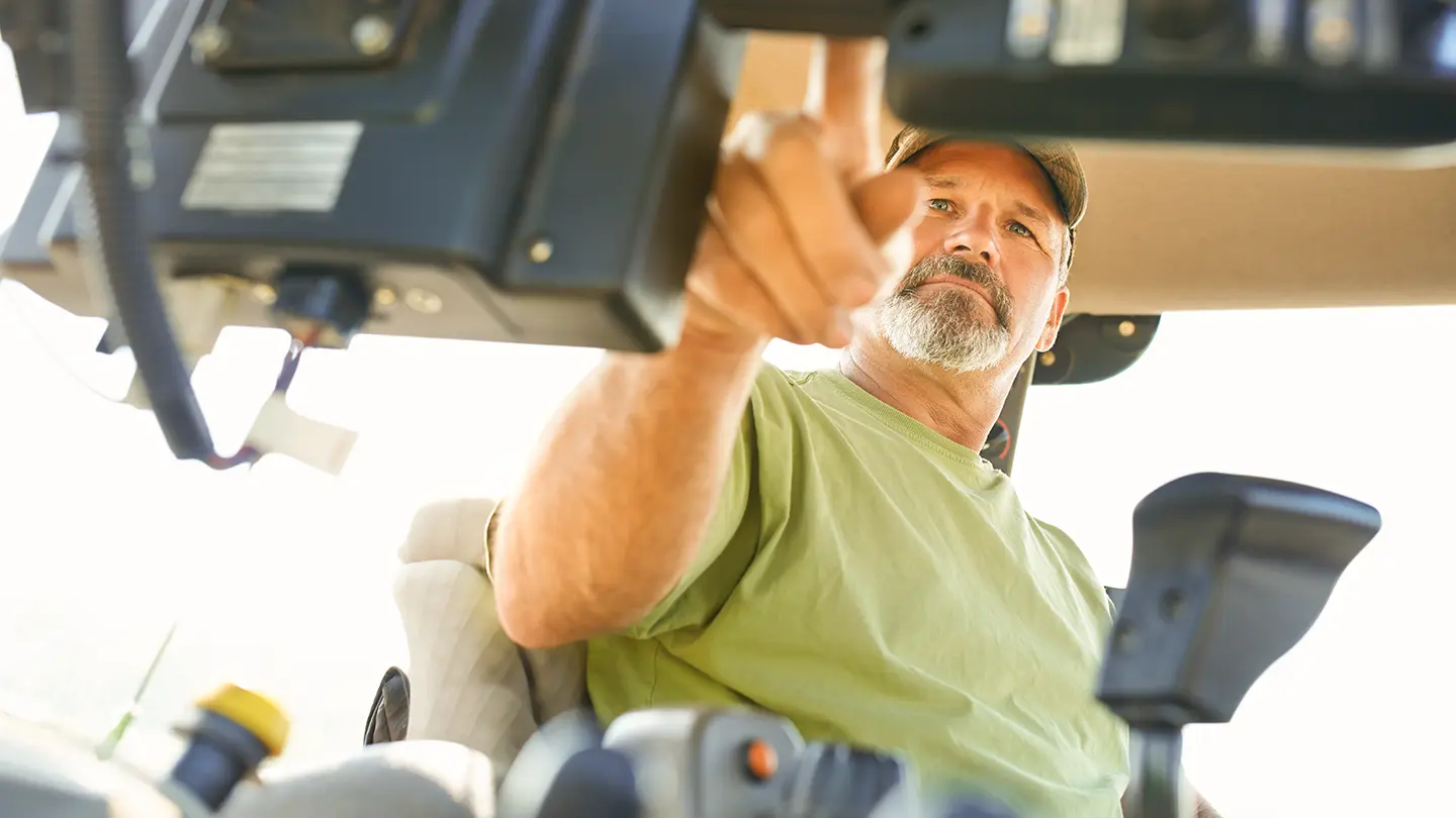 A close-up, low-angle shot of a middle-aged male farmer with a grey beard operating a touch-screen control panel inside a modern tractor cab.