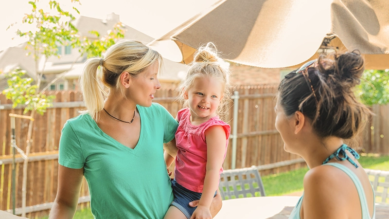 Two women chatting outdoors in a backyard setting while one holds a smiling young girl in a pink shirt.