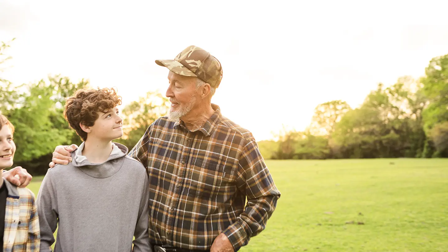 An older man in a camouflage cap and flannel shirt smiling warmly at a teenage boy in a green field during sunset.