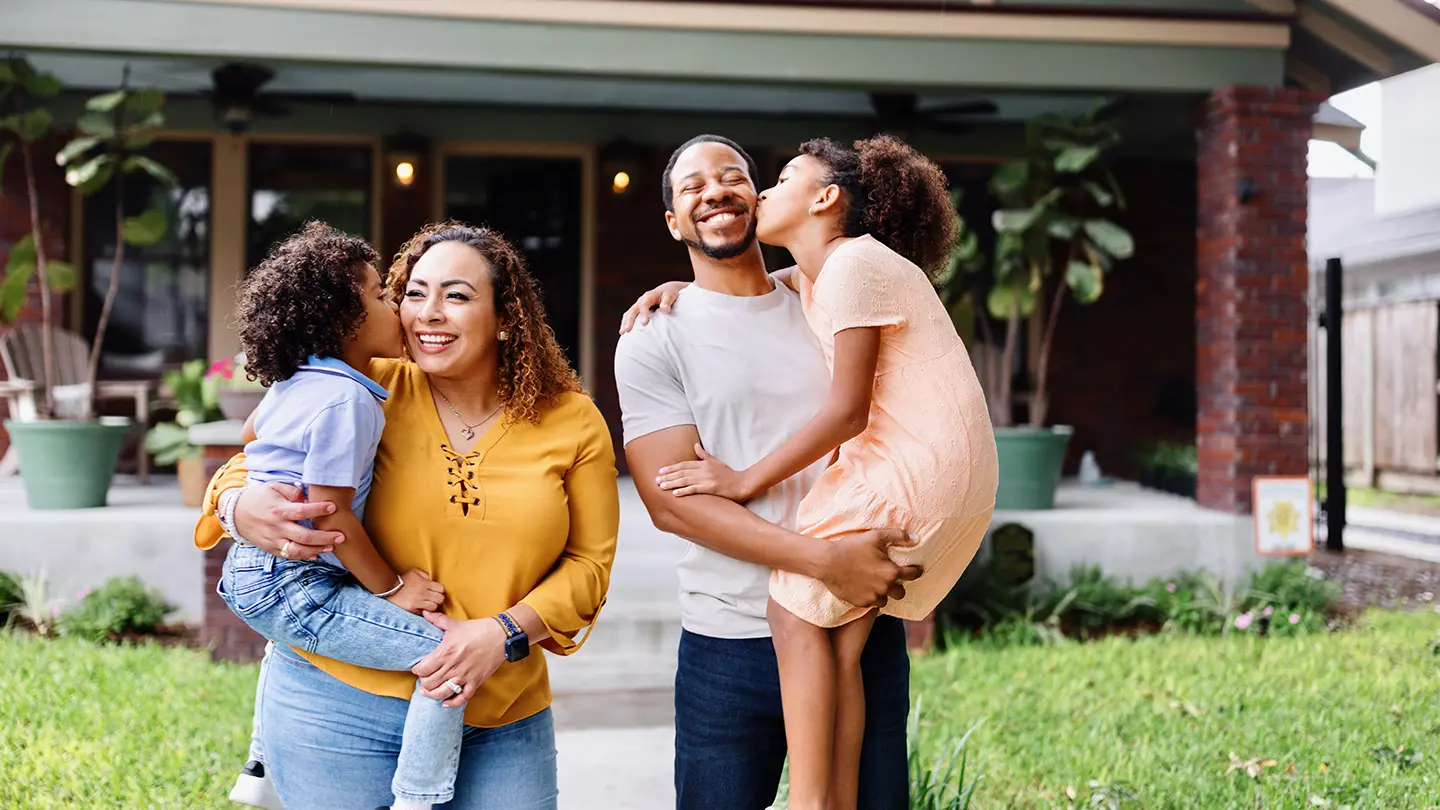 A happy young family of four standing in front of their home; the children are kissing their parents on the cheeks.