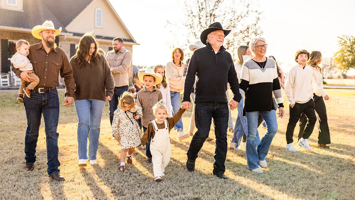 A wide shot of a large family group, including children in cowboy hats, walking across a lawn in front of a suburban house.