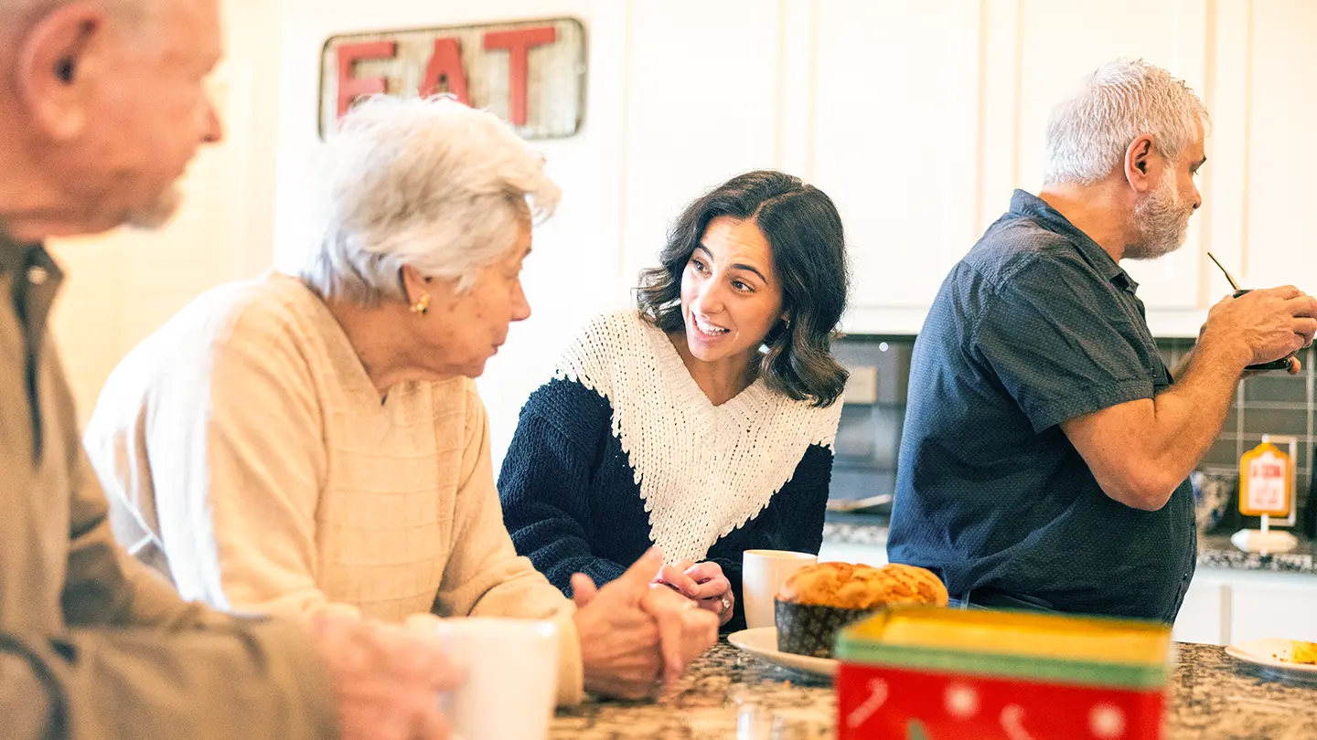 A young woman smiling and talking with older family members around a kitchen counter during a casual morning gathering with coffee.