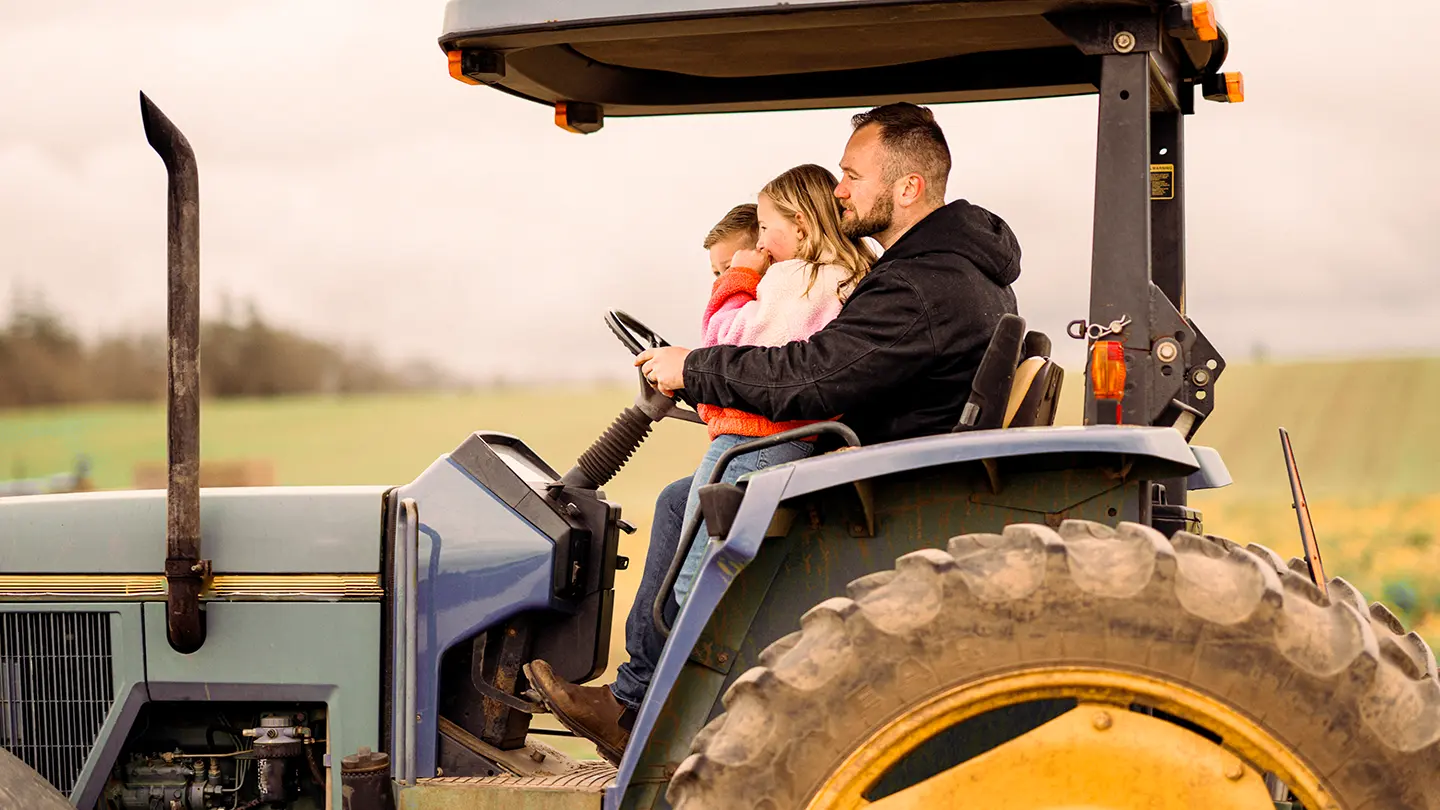 A man sitting on a blue tractor with two young children, showing them the controls in an open field.