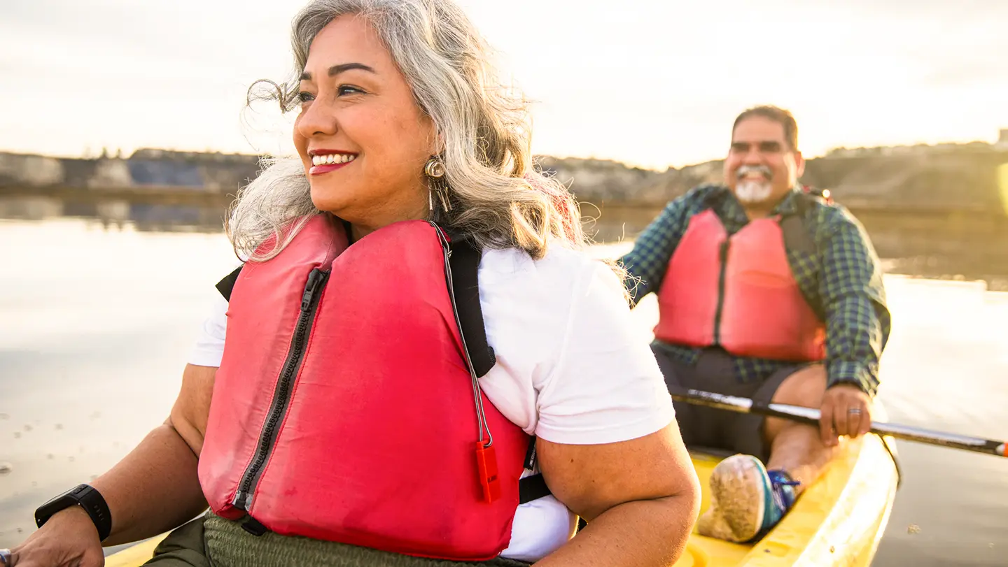 A smiling woman with grey hair wearing a red life vest while kayaking on a calm body of water at sunset.
