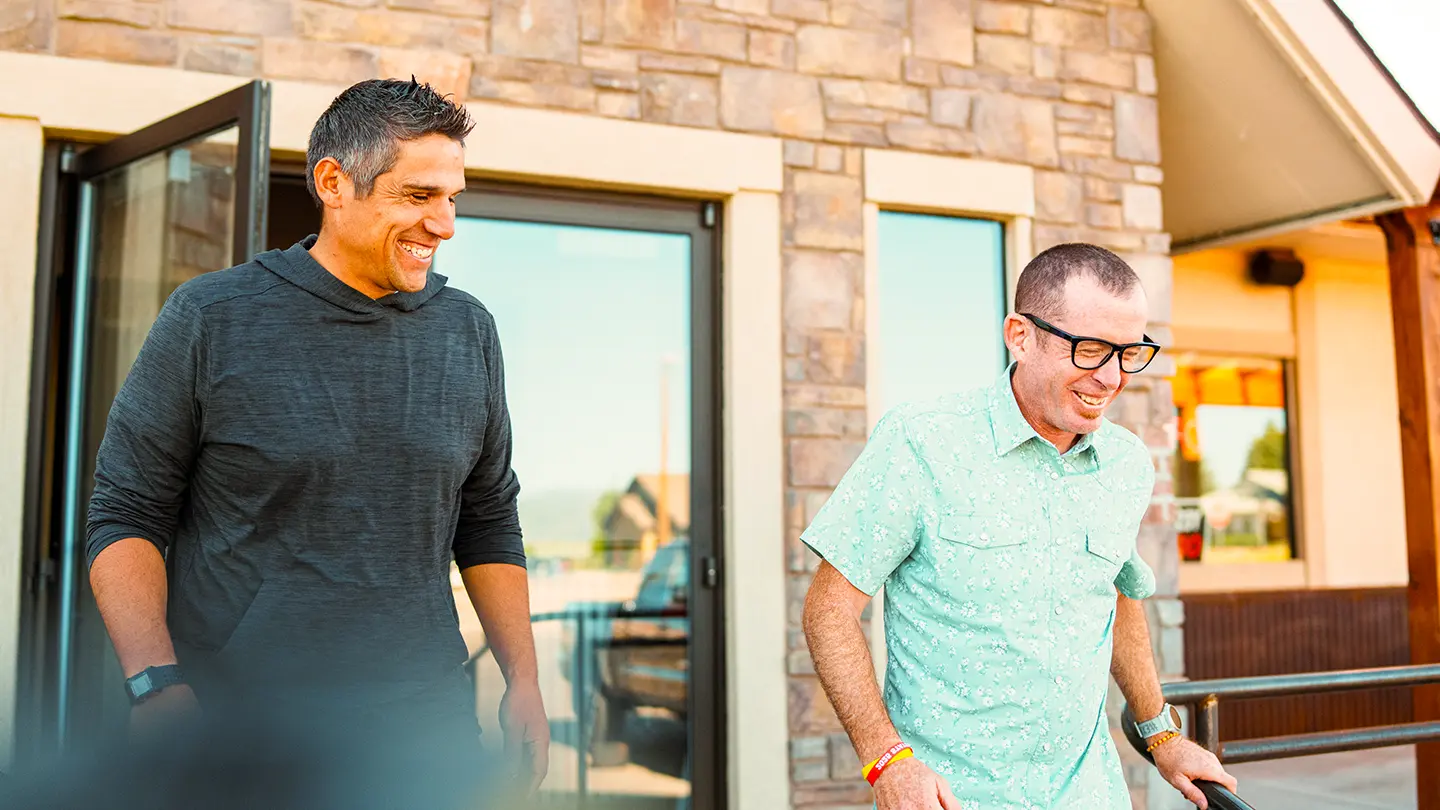 Two men laughing and talking as they walk out of a stone-faced commercial building.
