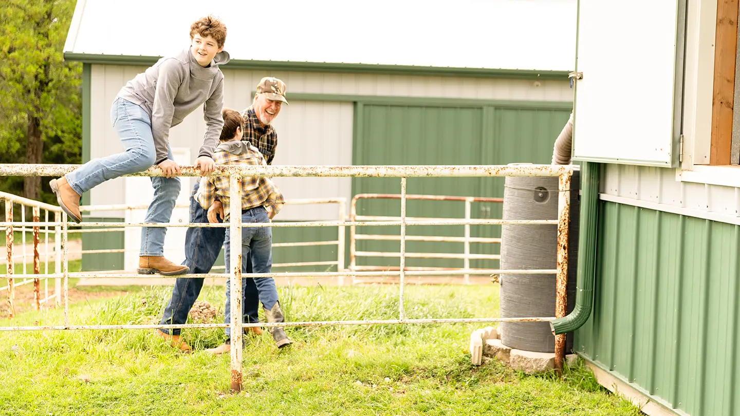 A group of young boys and an older man laughing while climbing over a white metal farm gate in front of a green barn.