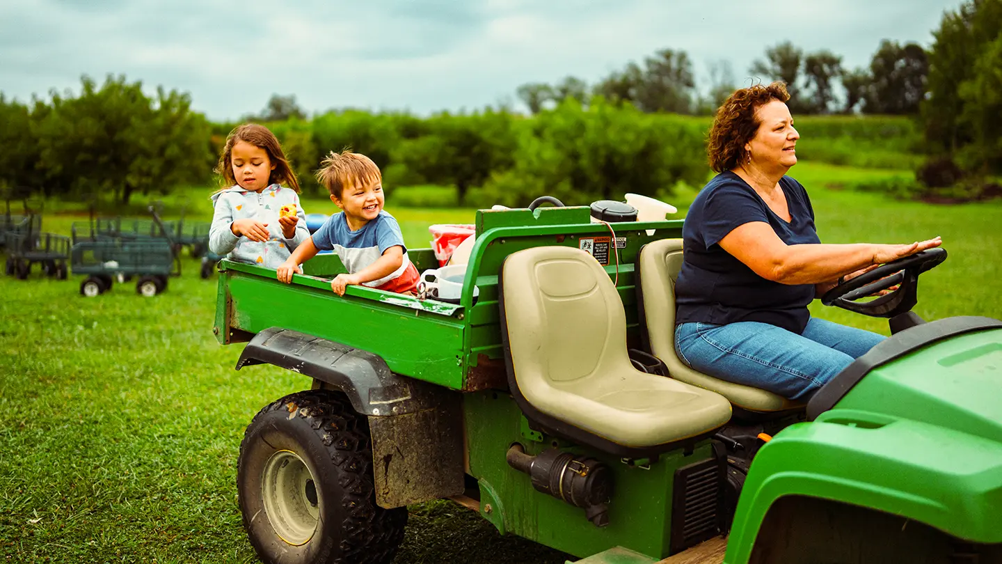 A woman driving a green utility vehicle through a grassy field with two happy young children riding in the back.