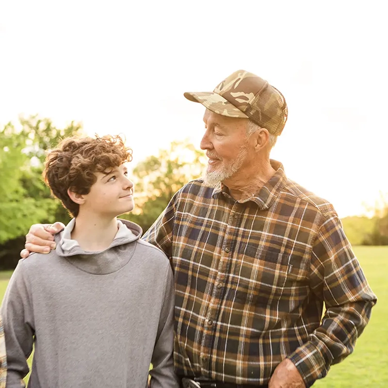 An older man in a camouflage hat and plaid shirt with his arm around a teenage boy with curly hair, both smiling at each other in a sunny field.