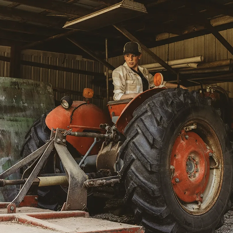 A woman wearing a plaid shirt and a baseball cap sitting on a large, vintage red tractor inside a rustic barn or shed.