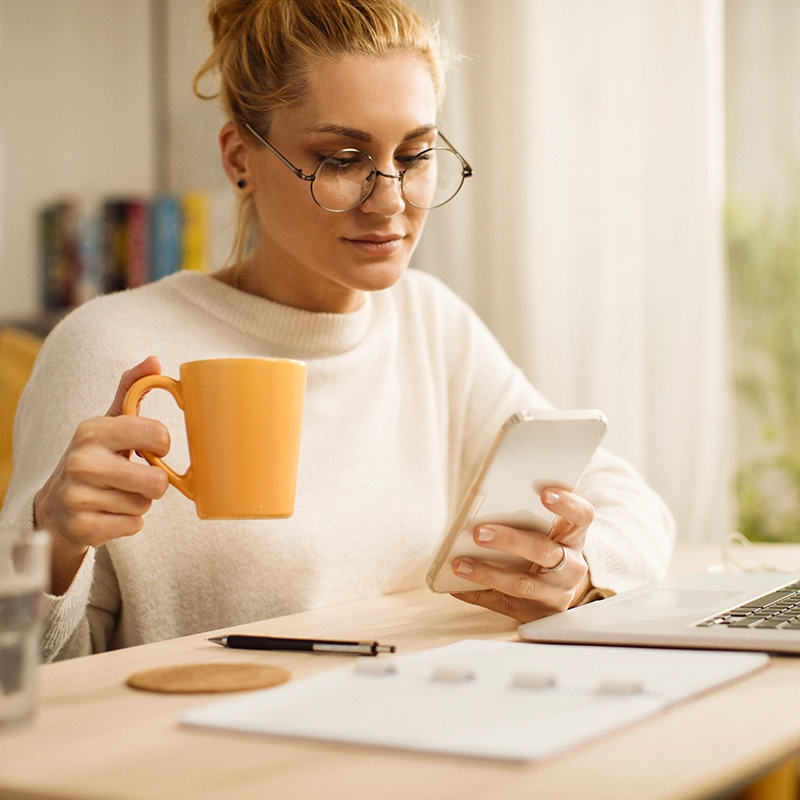 A woman with glasses and her hair in a bun sitting at a desk, holding a yellow mug and looking at her smartphone.