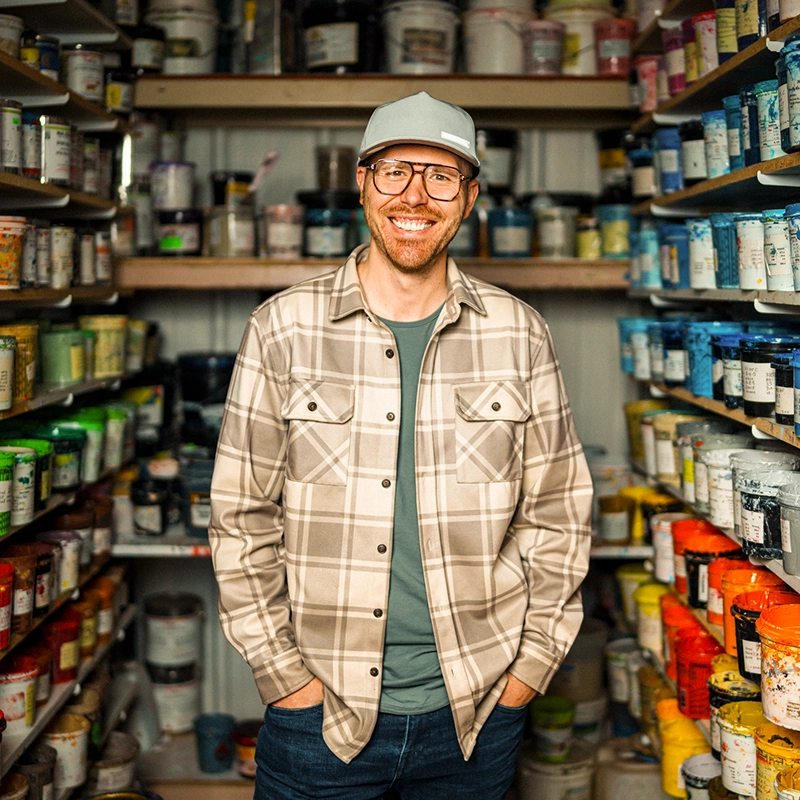 A smiling man in a plaid flannel shirt and grey baseball cap standing in a room filled with shelves of colorful paint cans.