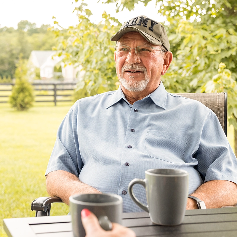An older man wearing an "ARMY" veteran cap sitting outdoors with mugs of coffee.