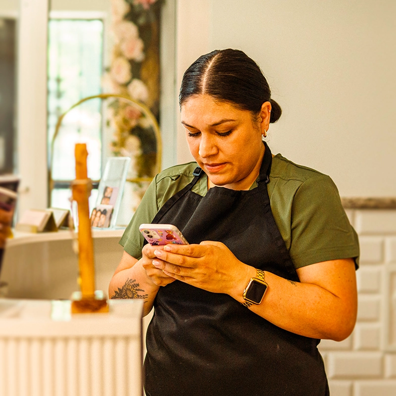 A woman in a black apron and green shirt looking down at her smartphone in a shop or cafe.