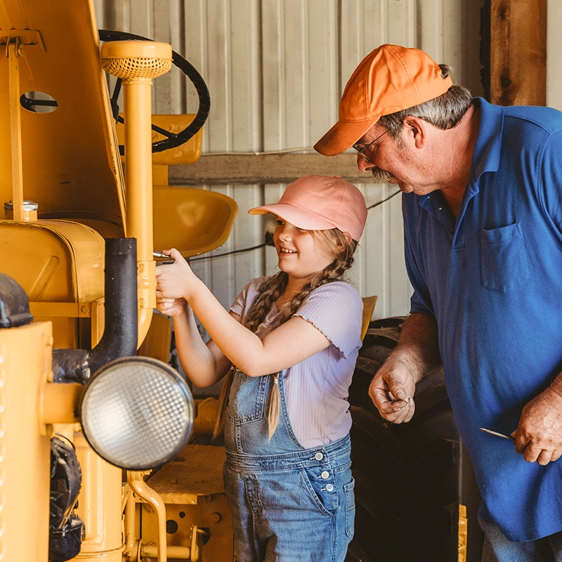 A man in an orange cap showing a young girl in overalls how to work on a yellow tractor.