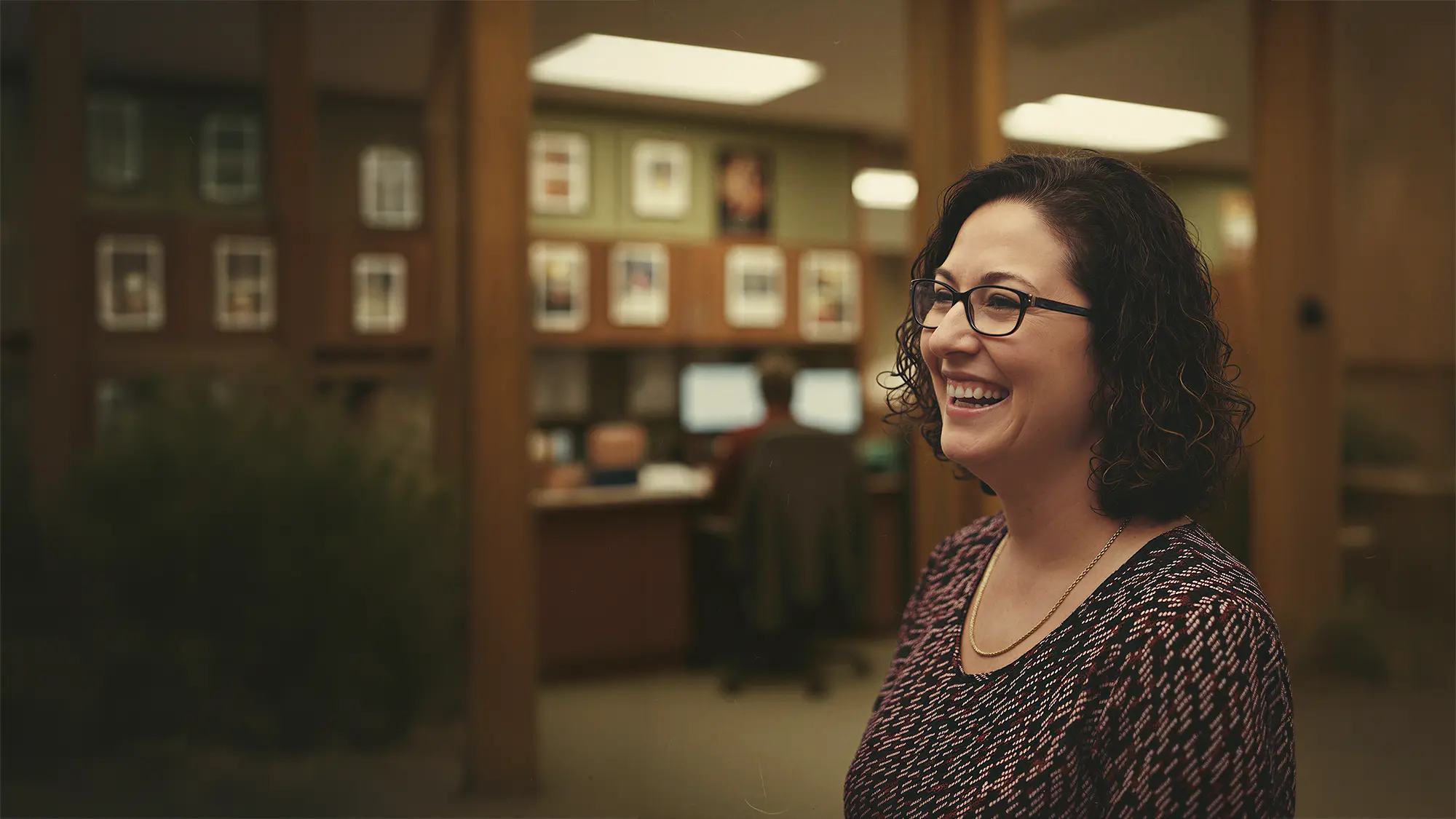 A woman with curly hair and glasses smiling warmly in a professional office environment with wooden pillars and workstations in the background.